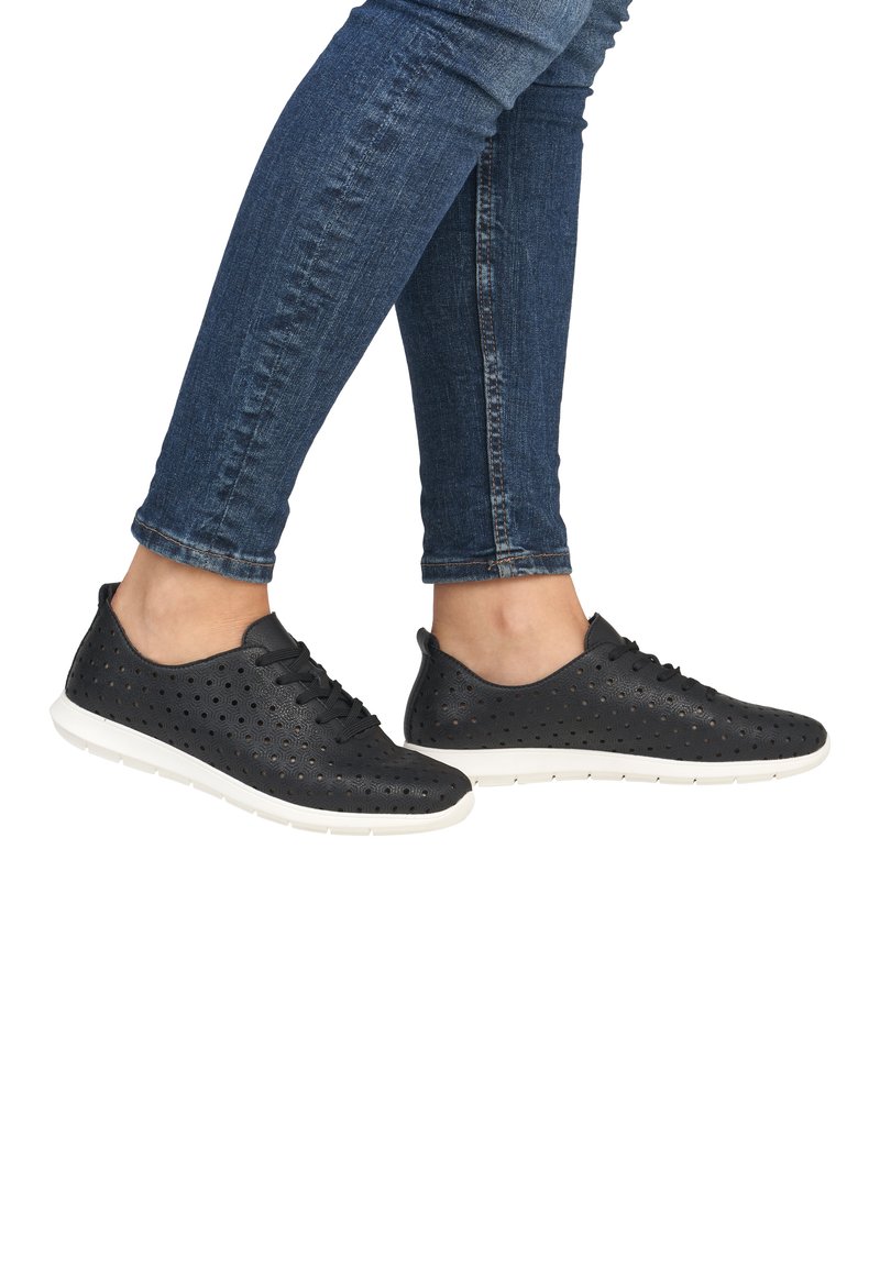 Black perforated sneakers with flat white soles; features laces, rounded toes, and a breathable design. Worn with fitted blue jeans.