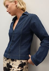 Camisa oscura de mezclilla con botones, cuello, detalles de costura horizontales y mangas largas; combinada con pantalones crema con un patrón negro.