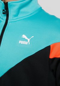 Chaqueta Puma con media cremallera, fabricada en suave tela turquesa con detalles en negro y naranja, y un logo blanco en el pecho.