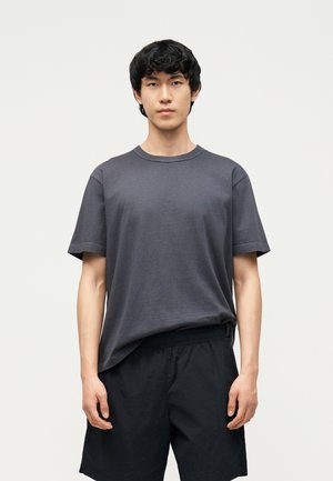 Junger Mann mit kurzen schwarzen Haaren, der ein dunkelgraues T-Shirt und schwarze Shorts trägt, steht vor einem schlichten hellen Hintergrund.