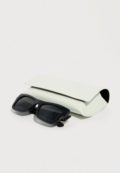 Μαύρα ορθογώνια γυαλιά ηλίου τοποθετημένα μπροστά από μια λευκή μαλακή θήκη σε απλό λευκό φόντο.