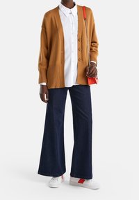 Senapsgul kofta med ribbat mönster, vit skjorta med knappar, vida mörka denimjeans och vita sneakers med röda accenter.