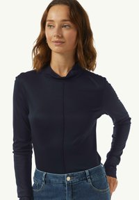 Marineblaues Langarmoberteil mit Stehkragen, das eine figurbetonte Passform und eine glatte Textur bietet. Kombiniert mit hellblauen Jeans.