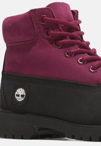Botas de ante burdeos con suela de goma negra. Presentan cordones redondos, ojales de metal y un logo circular con un diseño de árbol. Detalles de acento texturizado.