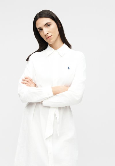 Femeie cu păr lung și închis la culoare, purtând o rochie albă cu guler și curea, cu brațele încrucișate, privind cu capul ușor înclinat pe un fundal simplu.