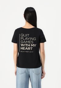 T-shirt en coton noir à manches courtes, avec un texte beige au dos : "ARRÊTE DE JOUER AVEC MON CŒUR BACKSTREET BOYS."