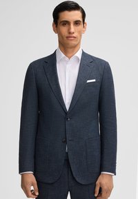 Giacca da abito blu navy realizzata in tessuto testurizzato, con due tasche frontali, rever a punte e due bottoni. Indossata sopra una camicia bianca.