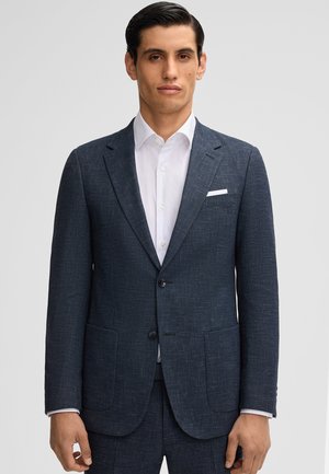 Giacca da abito blu navy realizzata in tessuto testurizzato, con due tasche frontali, rever a punte e due bottoni. Indossata sopra una camicia bianca.