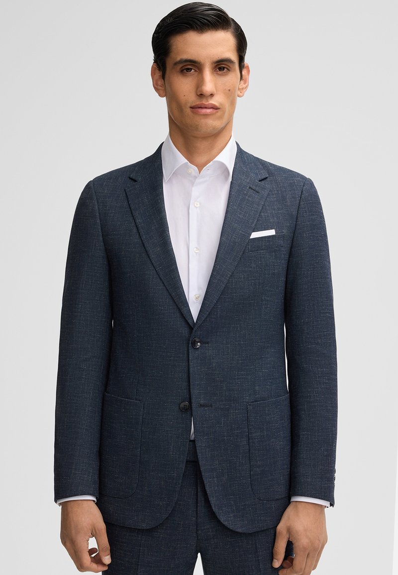 Giacca da abito blu navy realizzata in tessuto testurizzato, con due tasche frontali, rever a punte e due bottoni. Indossata sopra una camicia bianca.