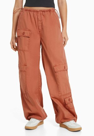 Pantalon cargo ample couleur rouille avec plusieurs poches à rabat, porté avec des baskets blanches et un haut court noir.