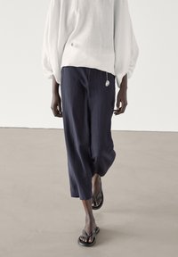 Top bianco in lino oversize con maniche a sbuffo, abbinato a pantaloni blu a tre quarti. Sandali neri dal design semplice completano il look.