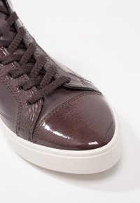 Gros plan d'une basket en cuir marron foncé avec un embout brillant texturé, des lacets assortis et une semelle en caoutchouc blanche sur fond blanc.