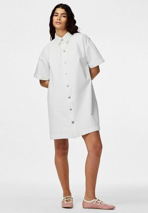 Nő áll kezekkel a hát mögött, fehér rövid ujjú gombos ingruhát és rózsaszín Mary Jane balerinacipőt visel fehér háttér előtt.