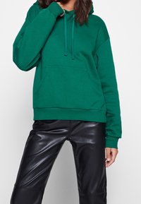 Grön huvtröja i mjukt material, med en framficka, justerbar snörning i huvan och ribbade mudd, i kombination med svarta läderbyxor.