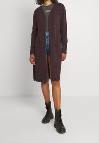 Kvinna som bär en lång mörk lila kofta, t-shirt med mörkgröna tryck, denimshorts och svarta snörskor, stående mot en enfärgad vägg.