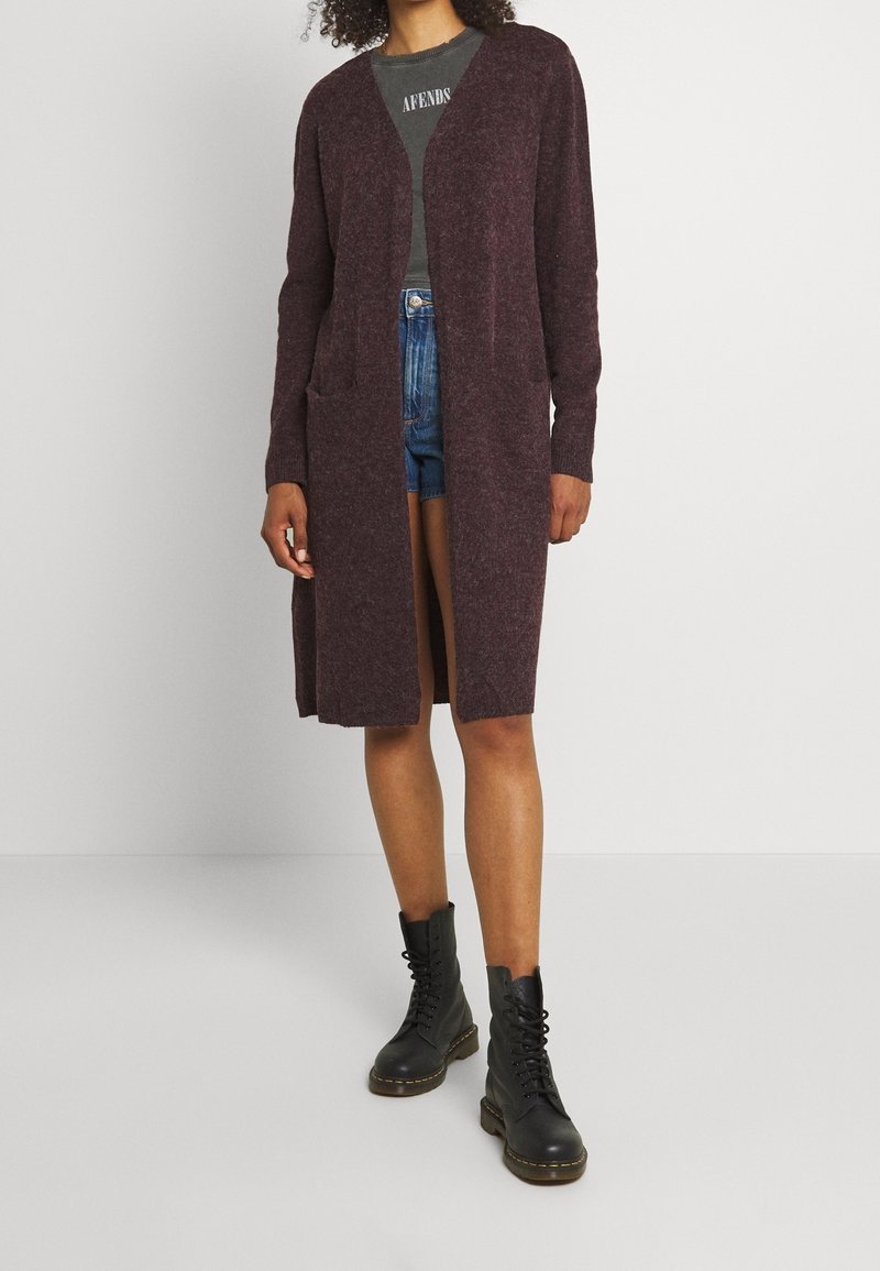 Kvinna som bär en lång mörk lila kofta, t-shirt med mörkgröna tryck, denimshorts och svarta snörskor, stående mot en enfärgad vägg.