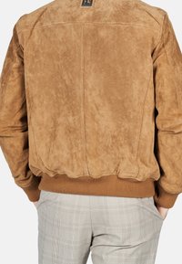 Achteraanzicht van een persoon die een bruine suède jas draagt met ribbelmanchetten en -zoom, gecombineerd met lichtgrijze geruite broek.