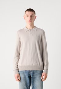 Ljusgrå långärmad polo med ribbad nederkant och ärmslut, tvåknappsknäppning och subtil branding på bröstet. Bärs med blåjeans.
