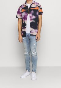 Hombre vistiendo una camisa de botones de manga corta con patrones en naranja, púrpura y negro, jeans claros rasgados, zapatillas blancas y una camiseta blanca.