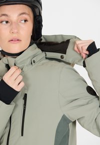 Giovane che regola il cappuccio di una giacca da esterno grigia chiara, indossando un casco nero, che guarda leggermente di lato.