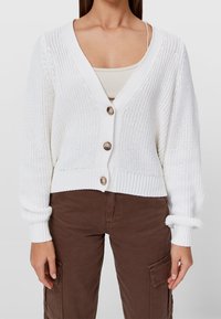 Bílý pletený kardigan s dlouhými rukávy, výstřihem do V a třemi velkými hnědými knoflíky. Žebrovaný lem a manžety, nošený přes béžový top.