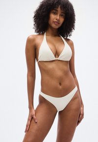 Μπικίνι εκρού χρώματος από υφασμα με υφή· τοπ σε σχήμα τριγώνου με λουρί γύρω από τον λαιμό· κάτω μέρος με χαμηλή μέση. Ομαλή επιφάνεια με μινιμαλιστικό σχεδιασμό.