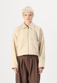 Beige zkrácená bunda vyrobená z nadýchané látky, s límcem, středovým zipem a dvěma předními kapsami. V kombinaci s hnědými skládacími kalhotami.