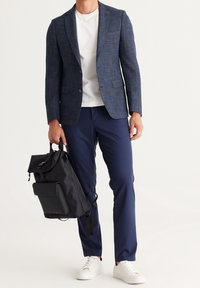 Muž v tmavě modrém saka, bílé košili, tmavě modrých kalhotách, bílých teniskách držící černý batoh za rukojeť na bílém pozadí.