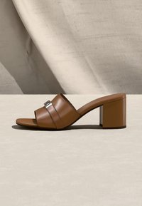 Καφέ δερμάτινο mule με ανοιχτό δάχτυλο και χοντρό τακούνι, διακοσμημένο με ασημένια αγκράφα, τοποθετημένο σε ουδέτερο φόντο και υφασμάτινο υπόβαθρο.