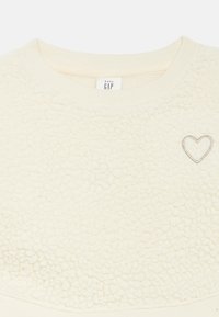 Φούτερ από βελούδινο φλις σε κρεμ απόχρωση με υφή και σχέδιο. Διαθέτει ριμπ λαιμόκοψη και μικρή ασημένια καρδιά στο μπροστινό μέρος.