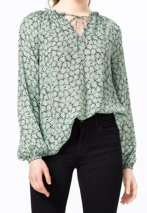 Femme portant un chemisier vert à manches longues avec motif de feuilles et un lien au niveau du col, associé à un pantalon noir ajusté.