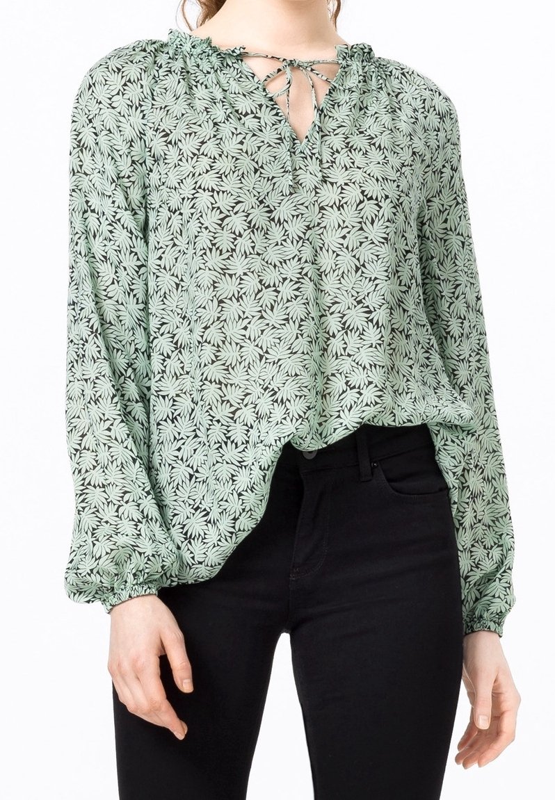 Femme portant un chemisier vert à manches longues avec motif de feuilles et un lien au niveau du col, associé à un pantalon noir ajusté.