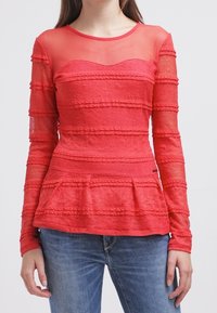 Top de manga larga rojo con escote de malla transparente, con estrías texturizadas en capas y cintura peplum acampanada. Combinado con denim.