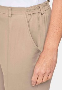 Beige broek met elastische tailleband, voorzien van plooien en zijzakken. Het materiaal lijkt glad met een lichte textuur.