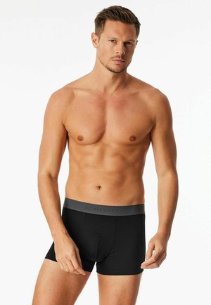 Man with short hair wearing black boxer briefs with a gray waistband, standing against a plain white background.