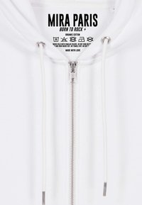 Hoodie blanc à zip avec cordons affichant l'étiquette "Mira Paris Born to Rock" et les instructions d'entretien à l'intérieur du col.