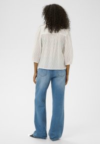 Personne aux cheveux bouclés portant une blouse blanche légère et un jean bleu ample, se tenant face à un fond blanc uni.