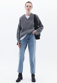 Harmaa neulepusero, jossa on V-aukko, yhdistettynä vaaleansinisiin suoriin farkkuihin ja mustiin nilkkasaappaisiin. Malli pitää mustaa olkalaukkua.