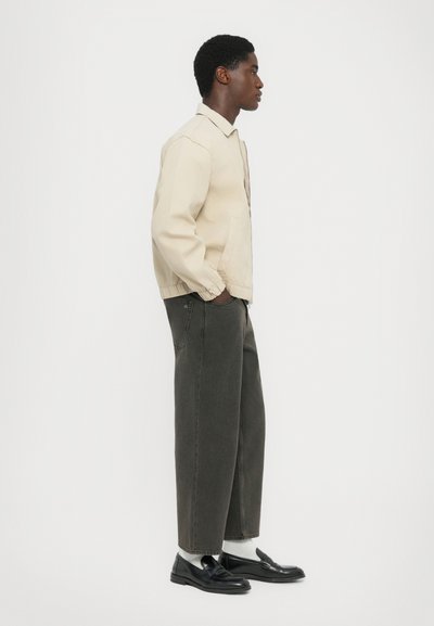 Ung man som bär en beige jacka, mörka vida jeans, vita strumpor och svarta loafers, stående i profil mot en vit bakgrund.