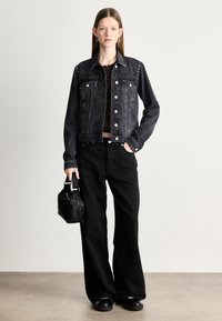 Musta farkkutakki, jossa on hopeiset niittikoristeet, yhdistettynä korkeavyötäröisiin mustiin housuihin ja pieneen mustaan käsilaukkuun. Sileä pinta ja slim fit -leikkaus.