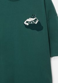 Ciemnozielona koszulka z grafiką przedstawiającą szczyt góry oraz czarny but turystyczny spoczywający na jej wierzchołku, umieszczona na górnej części klatki piersiowej.