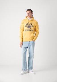 Sweat à capuche jaune avec un motif sur le devant et une poche kangourou, associé à un jean bleu clair et des baskets blanches. Le modèle se tient droit.