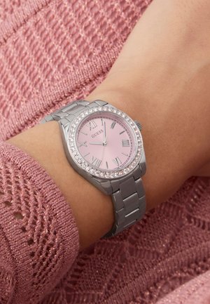 Montre-bracelet en métal argenté avec cadran rose, lunette en cristal, chiffres romains, portée au poignet sur un fond de pull rose texturé.
