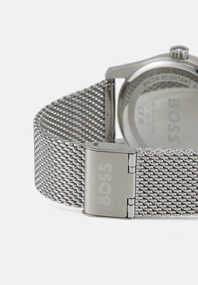 Montre avec bracelet en maille argentée et boîtier en acier inoxydable poli. Elle dispose d'une fermeture sécurisée et affiche un design minimaliste avec une résistance à l'eau.