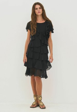 Pitkätukkainen nainen, jolla on polvipituinen musta kerroksittainen röyhelömekko ja kullanvärisillä niiteillä varustetut käärösandaalit, seisoo yksinkertaisen taustan edessä.