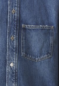Camicia di denim blu scuro, con colletto a punta, bottoni a scatto argento e taschino sul petto, con cuciture visibili e una texture morbida.