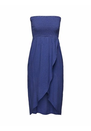 Osoba ubrana w jasnolirowy, oversize'owy sweatshirt, niebieską teksturowaną spódnicę z zakładkami, czarne sandały z odkrytymi palcami, stojąca na tle różowej ściany i białej podłogi.