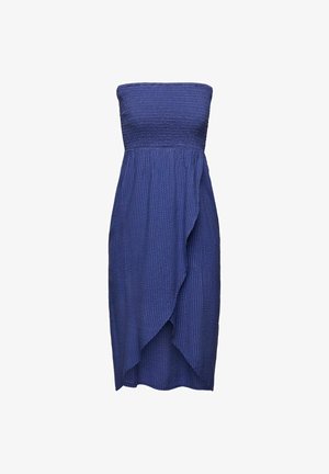 Personne portant un sweat-shirt ample vert clair, une jupe portefeuille bleue texturée, des sandales noires à bouts ouverts, debout contre un mur rose et un sol blanc.