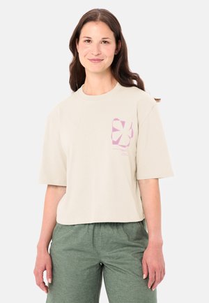 Femme portant une chemise beige à manches courtes avec un motif rose et un pantalon vert, debout devant un fond clair uni, souriant doucement.