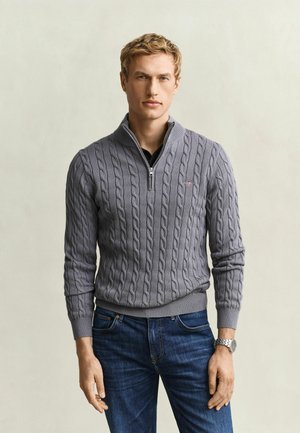 Homme blond portant un pull gris en maille torsadée avec fermeture éclair au quart, un jean bleu et une montre-bracelet argentée, debout devant un arrière-plan neutre.